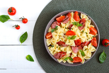 Tasty pasta farfalle with grilled sausages, fresh cherry tomatoes and basil on a plate on a white background. Top view, flat lay.