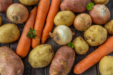 Young raw unpeeled potatoes, onions and carrots on dark wooden background. Mixed vegetables. Harvest concept. Flat lay composition. 