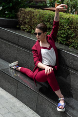 European girl sitting in the original pose in a business trouser suit in red. Women's jacket and pants cherry color on the Model with glasses with short hair.