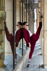 Fototapeta na wymiar Model girl with short hair. Brunette in sunglasses makes vertical twine near the glass shop window. The gymnast trains in an urban environment. Women's business jacket and burgundy pants.