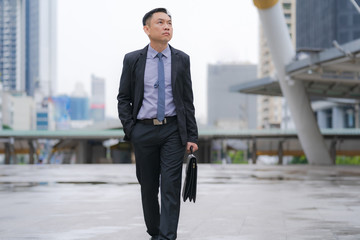 Asian Businessman walking and holding briefcase with business office buildings in the city...