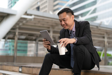 Fototapeta na wymiar Asian Businessman sitting and holding digital tablet with business office buildings in the city background