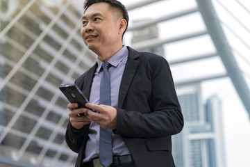 Asian Businessman standing and holding mobile phone with business office buildings in the city background