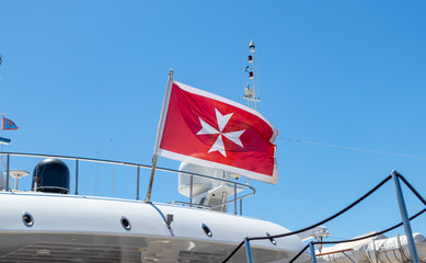 Maltese old flag on pole on ferry's stern. Ongoing cruise to islands. Blue sky background, close up view.