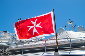 Maltese old flag on pole on ferry's stern. Ongoing cruise to islands. Blue sky background, close up...