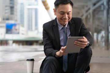 Asian Businessman sitting and holding digital tablet with business office buildings in the city background