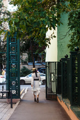 A girl in a white dress with polka dots and a hat walks against the background of a green house and a green gate turning her back