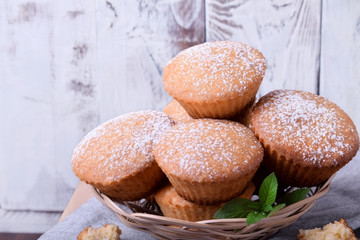 Fresh muffins topped with sugar powder in a wicker basket against the white wooden wall