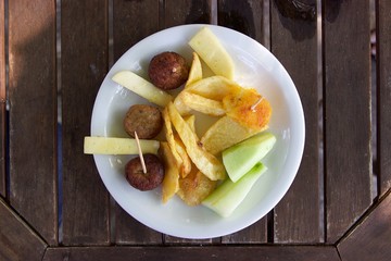 Top view Greek traditional meze food. A plate with appetizers of small meatballs, french fries, slices of cucumber and yellow cheese on a brown wooden table
