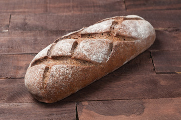 Whole loaf of dark homemade bread dusted with flour is lying on old brown boards