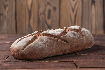 Whole loaf of dark homemade bread dusted with flour is lying on old brown boards