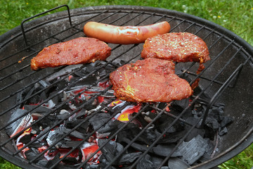 Steaks and sausage on round charcoal bbq. Glowing embers under a grill grid.