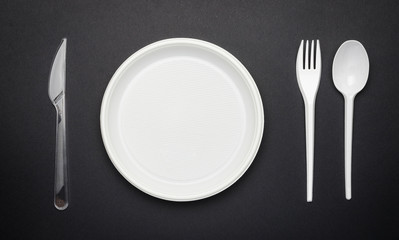 Disposable plastic tableware, fork, knife, spoon and plate on black background, top view