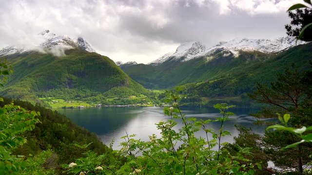 Spring Norway. Mountains on a fjord with snowy tops and pine forest. Water bay and dramatic grey cloudy sky. UHD