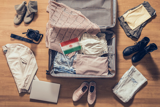 Woman's clothes, laptop, camera and flag of iran lying on the parquet floor near and in the open suitcase. Travel concept