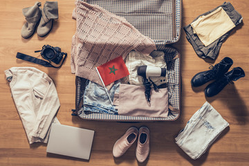 Woman's clothes, laptop, camera and flag of Morocco lying on the parquet floor near and in the open suitcase. Travel concept - obrazy, fototapety, plakaty