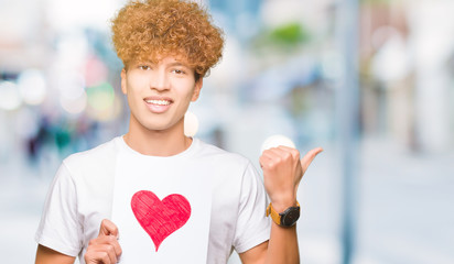 Young handsome man holding paper with red heart pointing and showing with thumb up to the side with happy face smiling