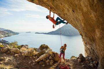 Young man starts climbing in cave, his female partner belaying him - obrazy, fototapety, plakaty
