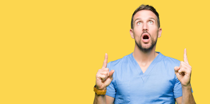 Handsome doctor man wearing medical uniform over isolated background amazed and surprised looking up and pointing with fingers and raised arms.