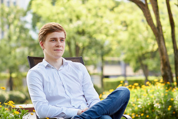 Young attractive man in white shirt sitting on brown chair in the park on green background - 277081576