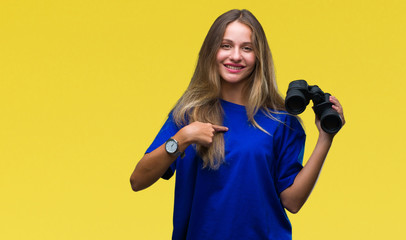 Young beautiful blonde woman looking through binoculars over isolated background with surprise face...