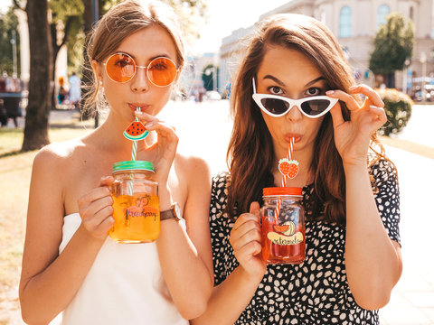 Two Young Beautiful Smiling Hipster Girls In Trendy Summer Clothes,dress.Sexy Carefree Women Posing In The Street.Positive Models Having Fun In Sunglasses.Drinking Fresh Cocktail Smoozy Drink