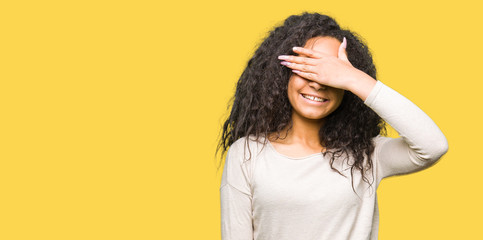 Young beautiful girl with curly hair wearing casual sweater smiling and laughing with hand on face covering eyes for surprise. Blind concept.