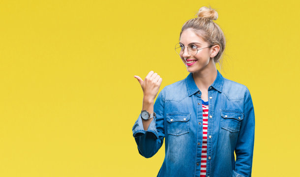 Young beautiful blonde woman wearing glasses over isolated background smiling with happy face looking and pointing to the side with thumb up.