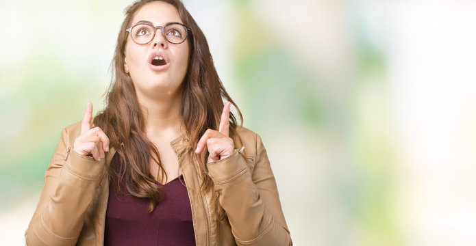 Beautiful plus size young woman wearing a dress and leather jacket over isolated background amazed and surprised looking up and pointing with fingers and raised arms.