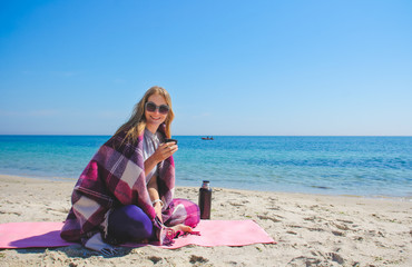 Fototapeta na wymiar Blonde girl sitting on the beach with thermos and drinking tea. Young smiling woman wrapped in warm pink blanket near sea.