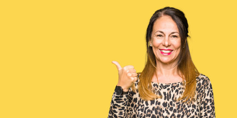 Beautiful middle age woman wearing leopard animal print dress smiling with happy face looking and pointing to the side with thumb up.