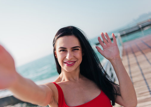 Portrait of young smiling woman in a red swimsuit takes a selfie on the beach