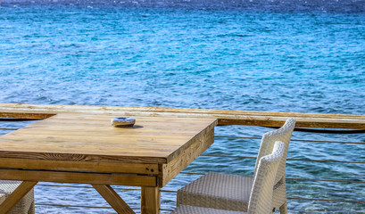 Greece promenade waterfront resort district outdoor patio cafe exterior wooden furniture of white chair and ashtray on brown wooden table with vivid blue Mediterranean sea background, copy space  