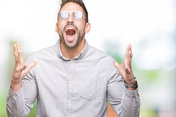 Naklejka premium Young handsome man wearing sunglasses over isolated background crazy and mad shouting and yelling with aggressive expression and arms raised. Frustration concept.