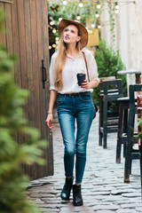 Young attractive woman in casual style clothes walking along the courtyard of the old city with a cup of coffee on the go