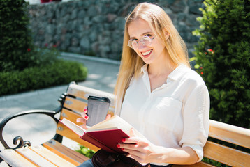 Young attractive blonde female student in glasses reading a book while sitting on a bench outdoors and looking into the camera