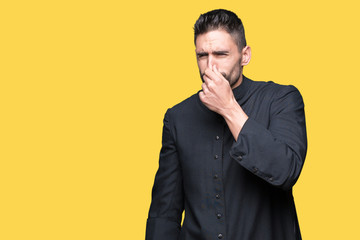 Young Christian priest over isolated background smelling something stinky and disgusting,...
