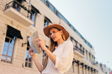 Young casual style tourist woman in felt hat looking into the city map against the background of old urban architecture. The concept of travel to unfamiliar cities