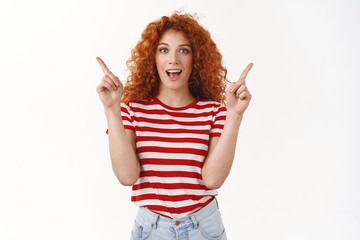 Wow check this out. Enthusiastic cheerful redhead attractive curly hair woman pointing up sideways left right choice promote product show advertisement thrilled turn attention, white background