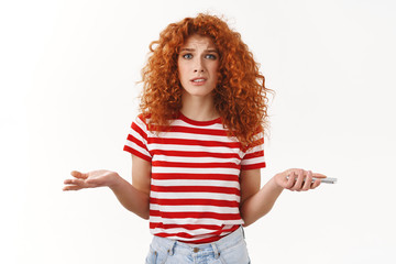 Obraz premium Confused uncertain stylish beautiful redhead curly girlfriend shrugging dismay puzzled holding smartphone hands spread sideways stare questioned trying understand who spam mailbox