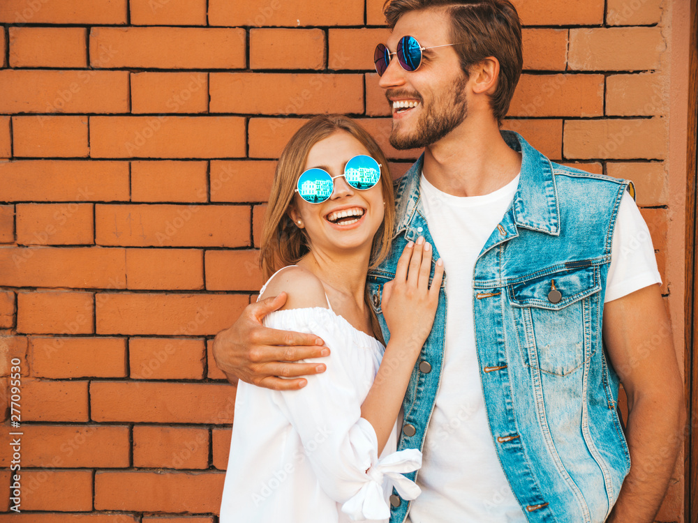 Wall mural Smiling beautiful girl and her handsome boyfriend. Woman in casual summer dress.Man in jeans clothes.Happy cheerful family.Female having fun on the street near brick wall.Hugging couple in sunglasses