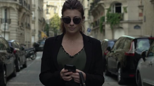 Attractive caucasian classy woman with sunglasses, freckles, piercings, black jacket and red hair writing a text message smiling walking through the street, during sunny day in Paris. Slow motion.