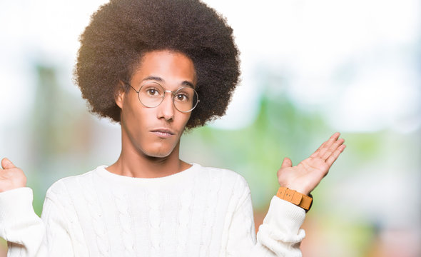 Young african american man with afro hair wearing glasses clueless and confused expression with arms and hands raised. Doubt concept.