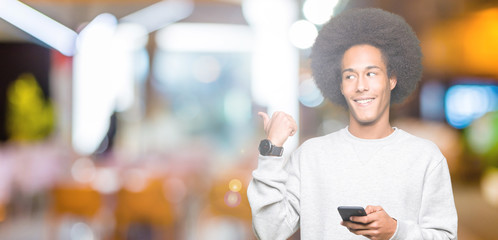 Young african american man with afro hair using a smartphone pointing and showing with thumb up to the side with happy face smiling