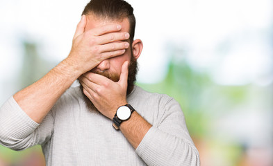 Young blond man wearing casual sweater Covering eyes and mouth with hands, surprised and shocked. Hiding emotion