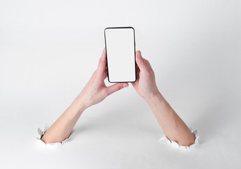 Female hands holding smartphone through the torn holes of white background. Creative art