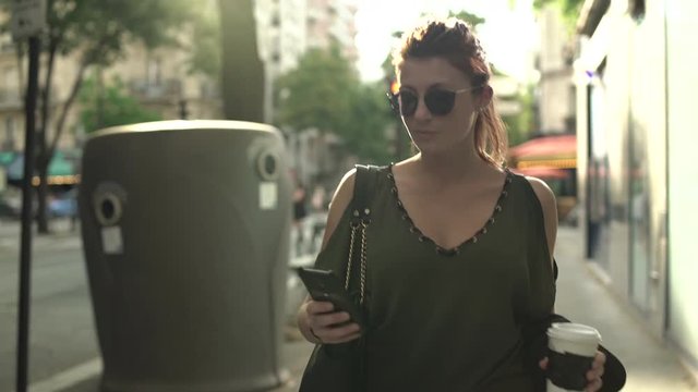 Attractive smiling caucasian woman with sunglasses, freckles, piercings and red hair writing text message to boyfriend on her smartphone and drinking coffee walking through the street in Paris. 4K.