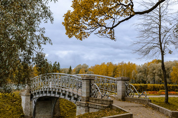 Openwork metal bridge in the autumn Park with a sandy path with trees covered with green, yellow and orange foliage against the cloudy sky.