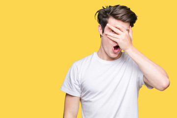 Fototapeta na wymiar Young handsome man wearing casual white t-shirt over isolated background peeking in shock covering face and eyes with hand, looking through fingers with embarrassed expression.