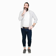 Fototapeta premium Young man with long hair, beard and earrings with hand on chin thinking about question, pensive expression. Smiling with thoughtful face. Doubt concept.